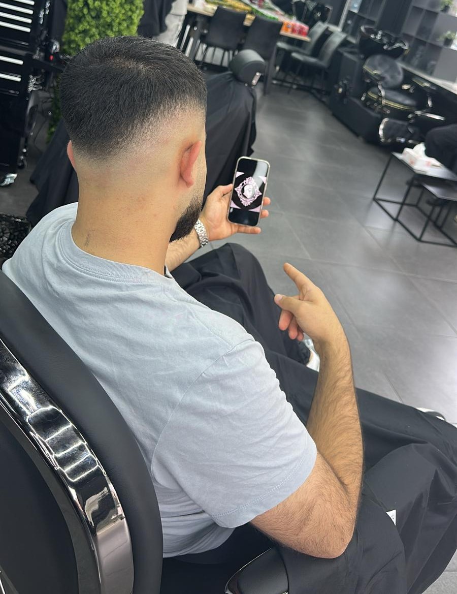 Een klant in de barberstoel bekijkt zijn verse skin fade op zijn telefoon, met de groene plantenwand achter hem
